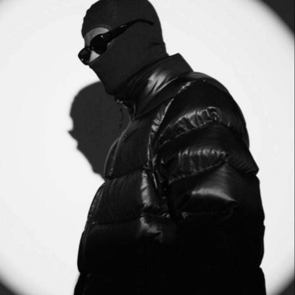 Person wearing a black puffer jacket and sunglasses against a white background