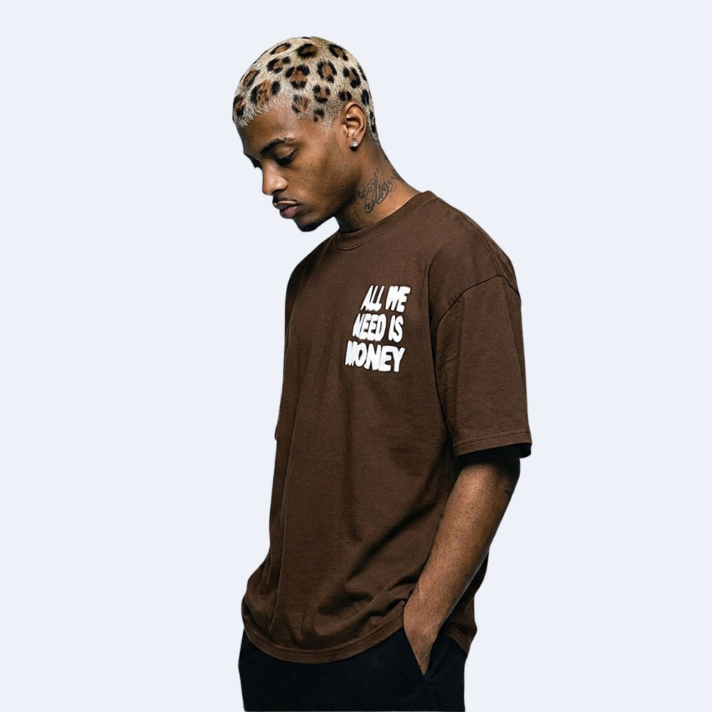 A man wearing a brown crewneck t-shirt with the text 'ALL WE NEED IS MONEY' printed in white on the chest, posed against a light gray background.