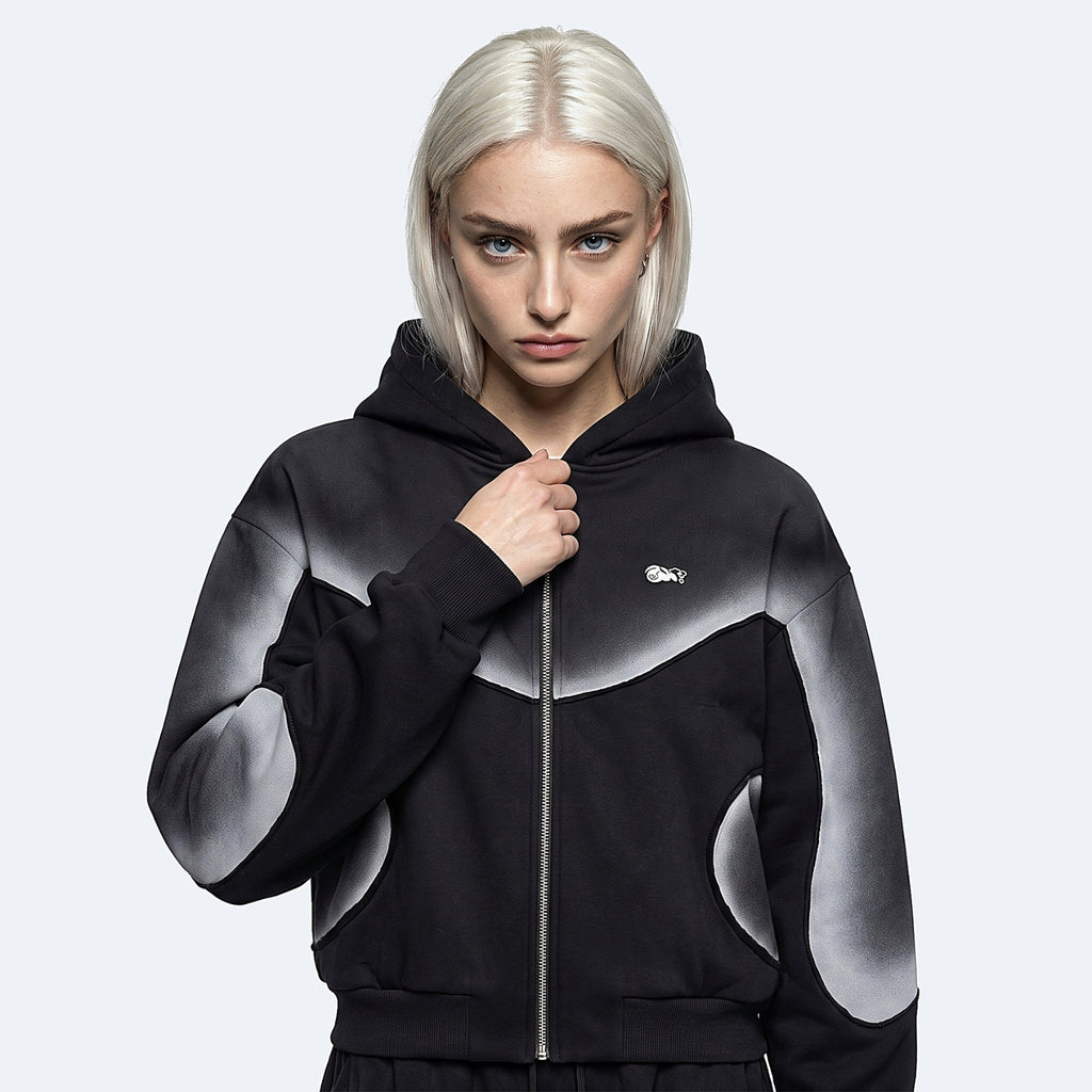 A model with platinum blonde hair wearing a black and gray gradient zip-up hoodie with a small white Lacoste logo on the chest, set against a light gray studio background.