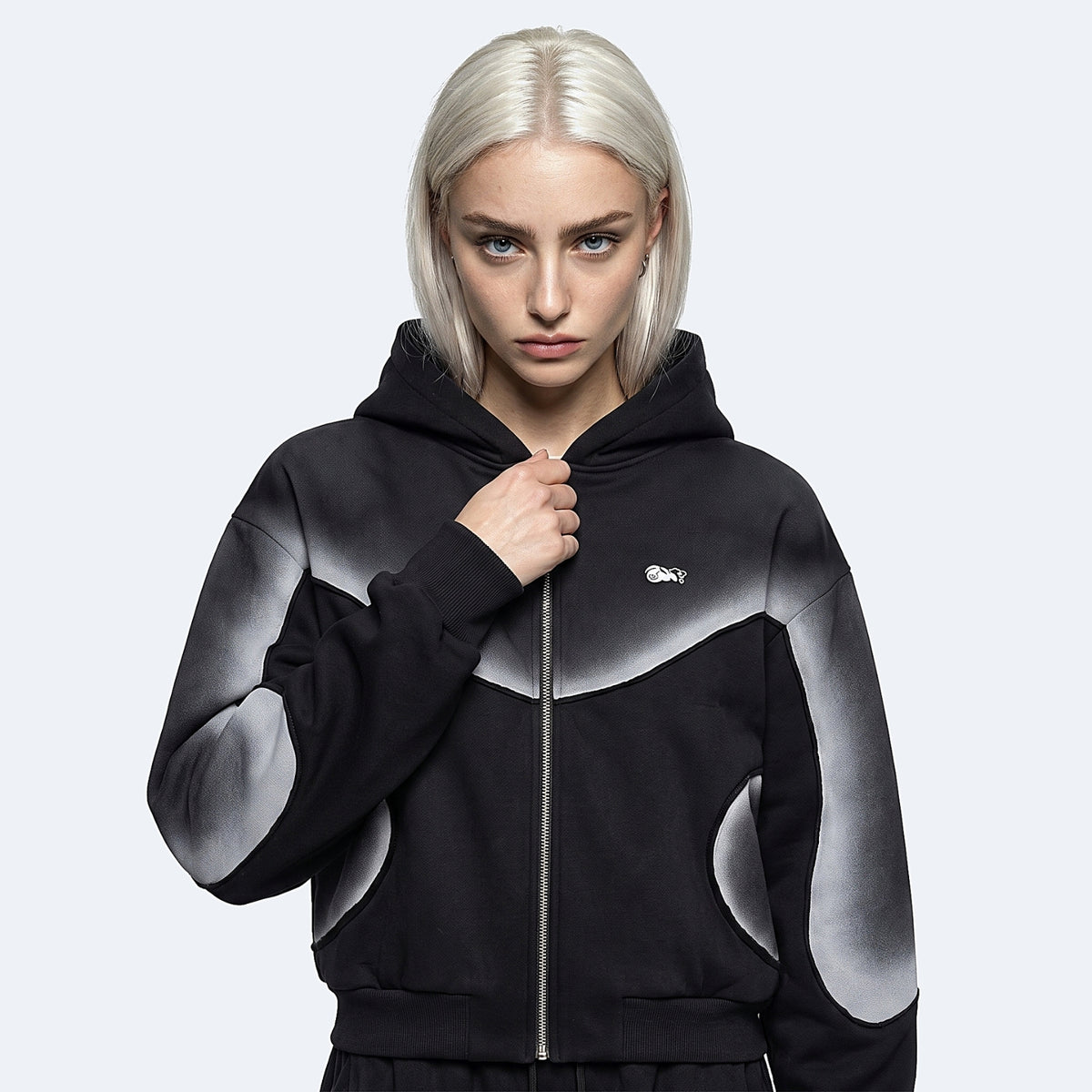 A model with platinum blonde hair wearing a black and gray gradient zip-up hoodie with a small white Lacoste logo on the chest, set against a light gray studio background.