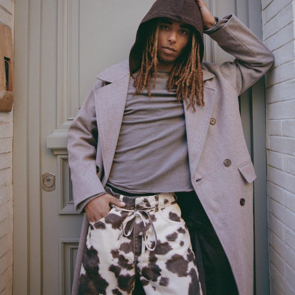 Person wearing a long coat over a gray shirt and cow print pants, standing against a door.