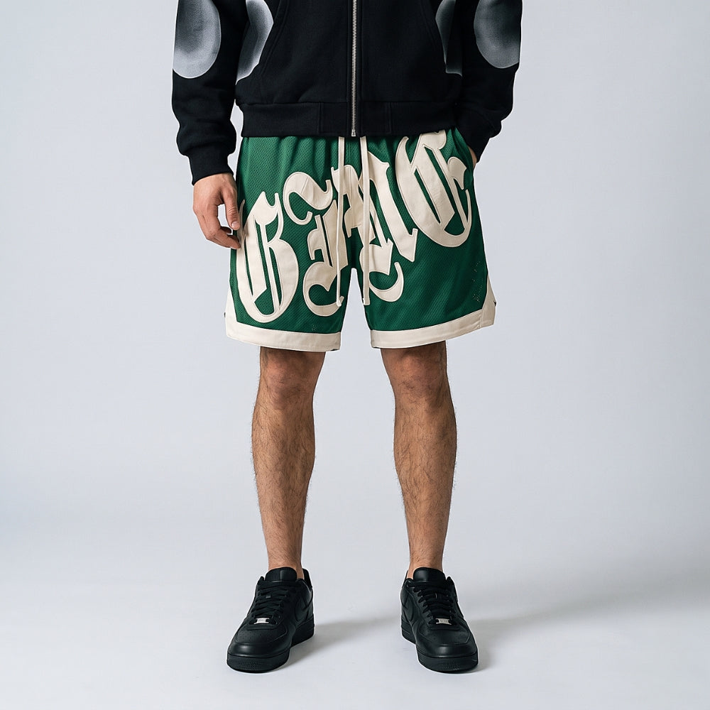 Green. A model wearing green athletic shorts with a large white script graphic across the front, paired with a black zip-up hoodie and black sneakers.