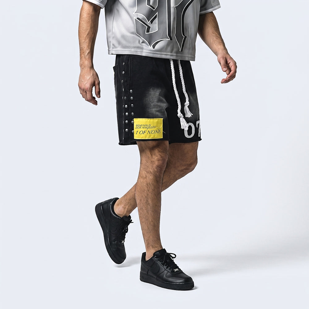 A model wearing black distressed athletic shorts with white drawstrings, a yellow graphic patch, and silver side grommets, paired with a gray graphic t-shirt and black sneakers, against a light gray studio background.