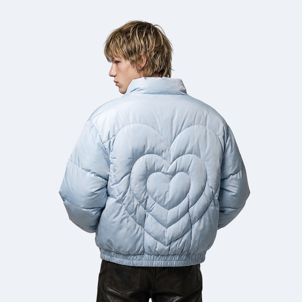 Blue. A model shown from the back wearing a light blue puffer jacket with a large quilted heart design on the back, paired with dark brown leather pants.