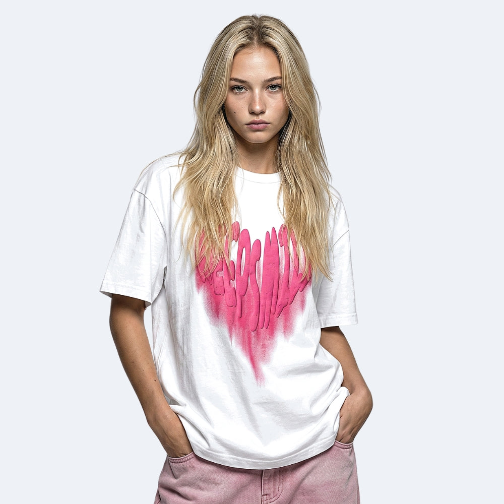 A blonde woman wearing a white oversized t-shirt with a pink dripping hand graphic on the front, paired with dusty pink pants, against a light gray background.