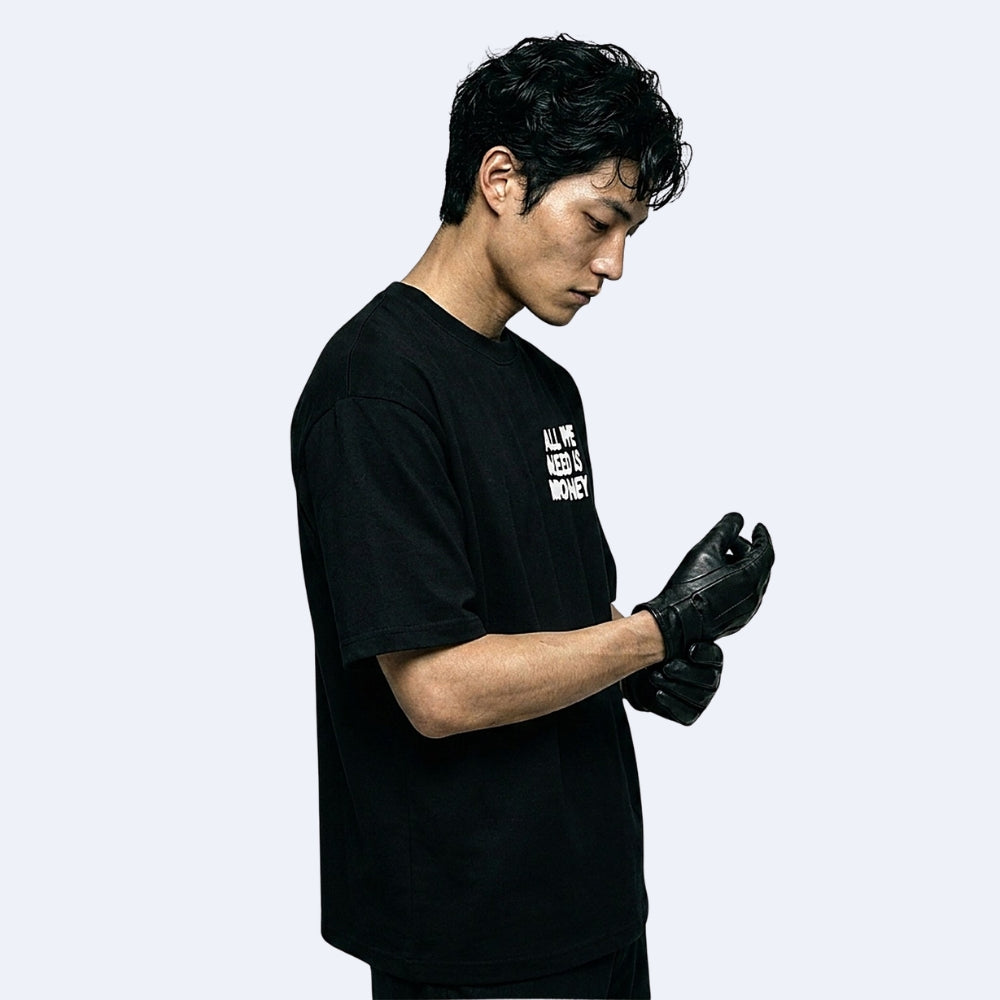 A man in a black t-shirt and black gloves looking down, set against a solid light gray background. The t-shirt features white text on the left chest that reads 'ALL WE NEED IS ONE WOMAN'.