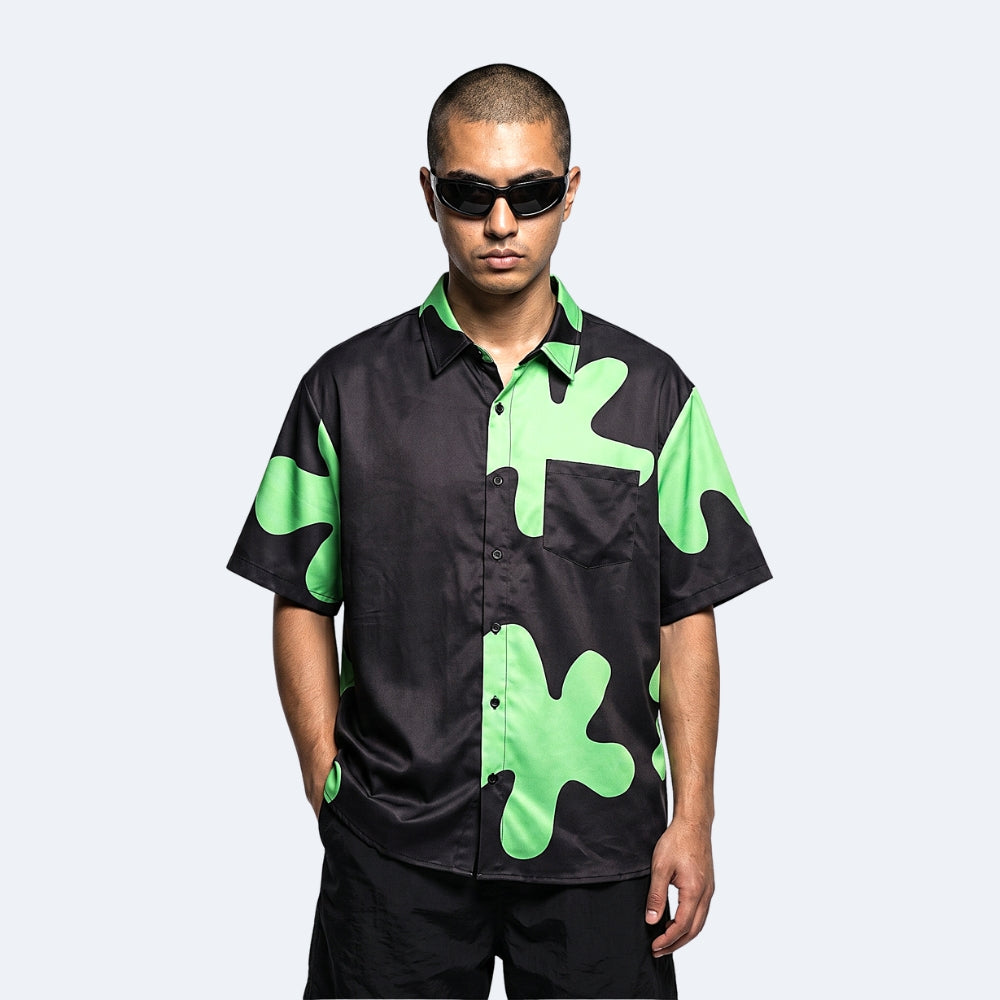 Black & Green. A man wearing a black short-sleeve button-up shirt with bright green abstract camouflage patterns, styled with black sunglasses and black pants.