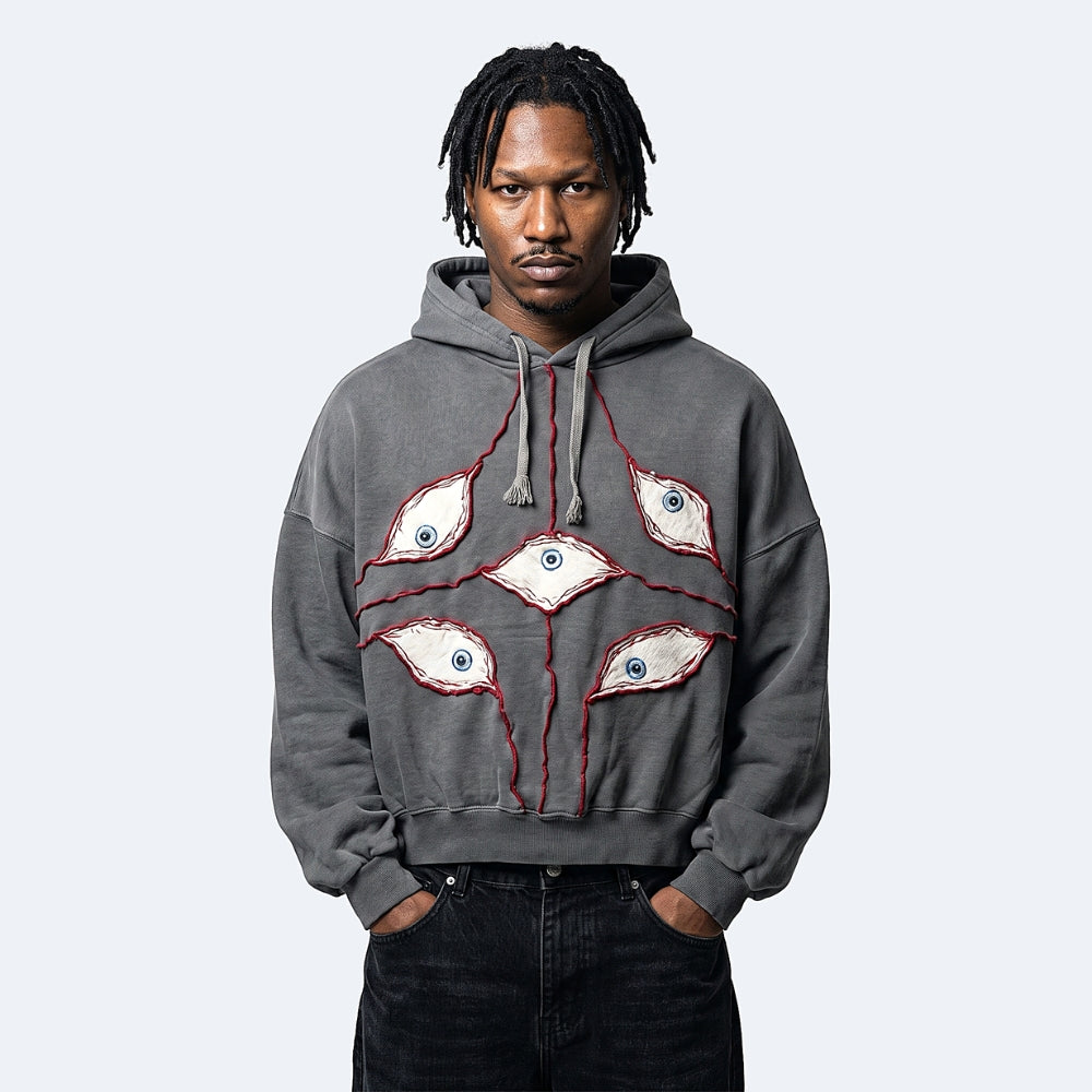 A man wearing a dark gray oversized hoodie with a unique graphic design of five embroidered eyes with red stitched outlines, paired with black jeans, against a light gray background.