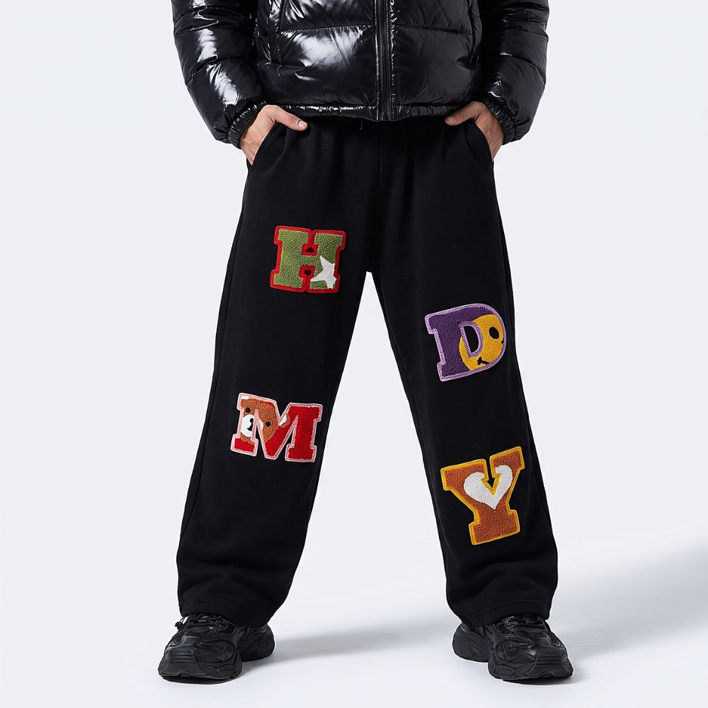 Black. Black sweatpants with colorful embroidered varsity letters on the legs, shown on a model wearing a black puffer jacket and black sneakers.