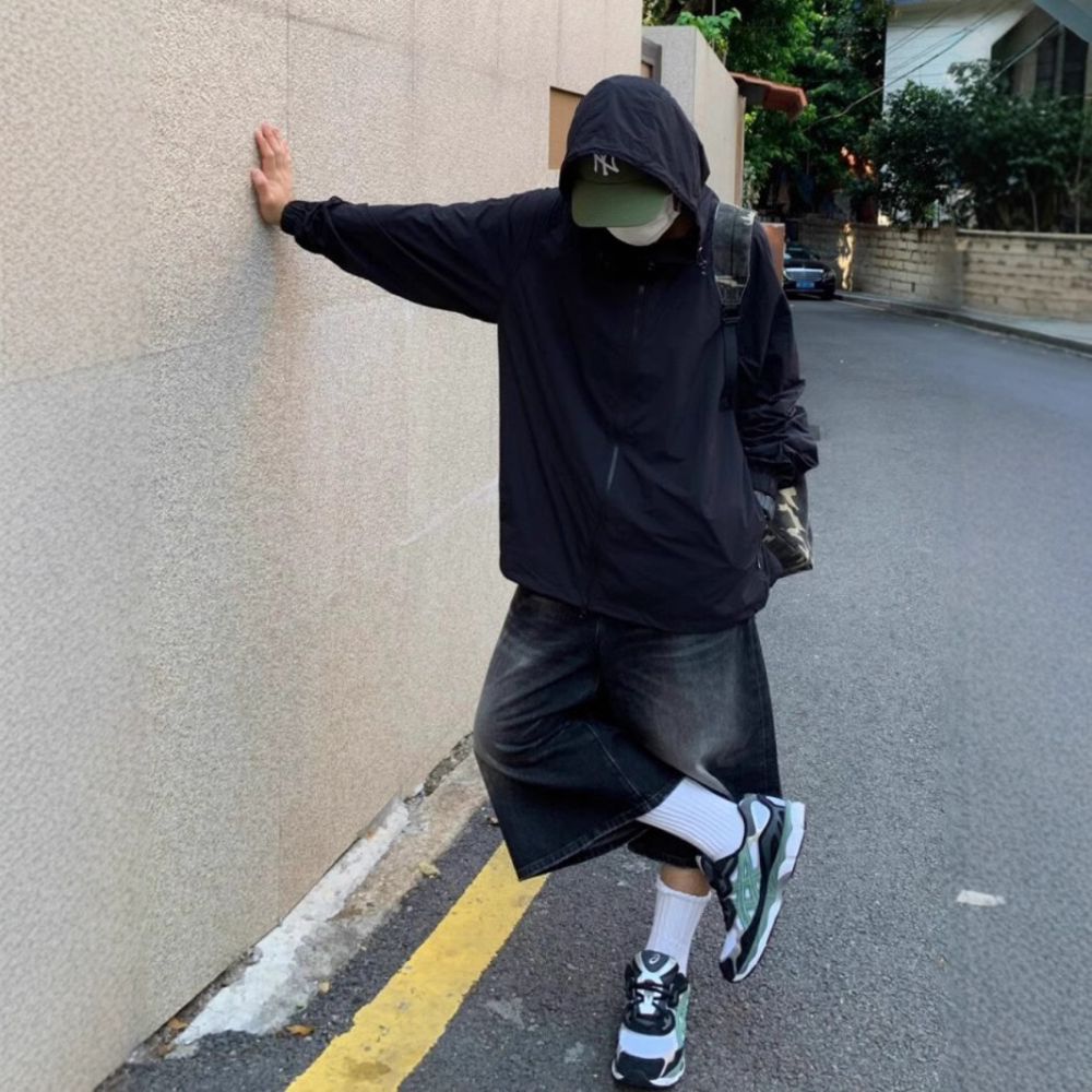 Person wearing a black hoodie and shorts leaning against a wall on a street.