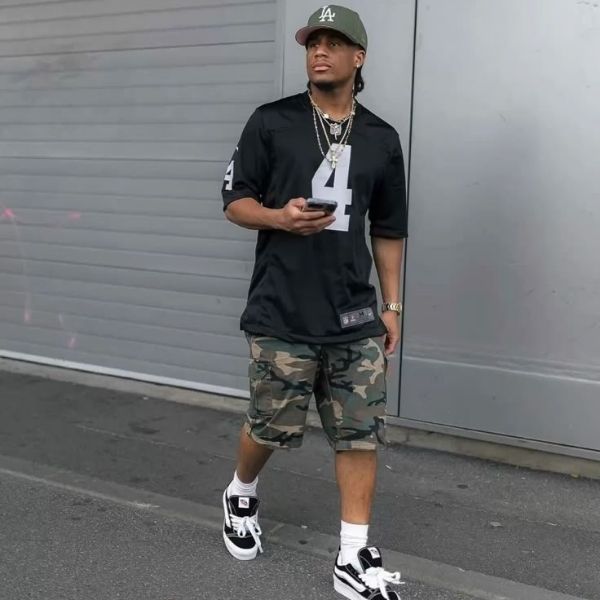 Man wearing a black sports jersey with number 4, camouflage shorts, and a cap, standing against a gray wall.
