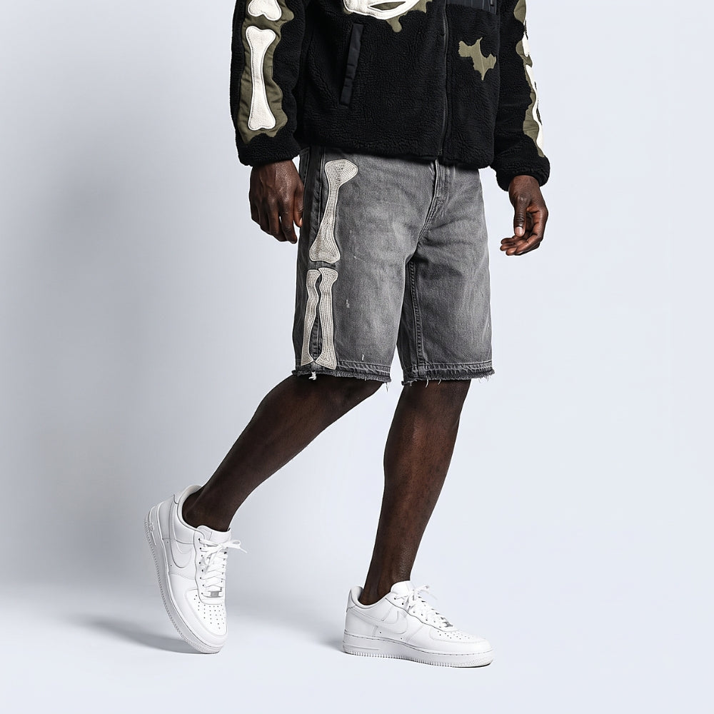 A model wearing gray denim shorts with a large white skeleton print down the side, styled with a black fleece zip-up hoodie and white sneakers against a light gray studio background.
