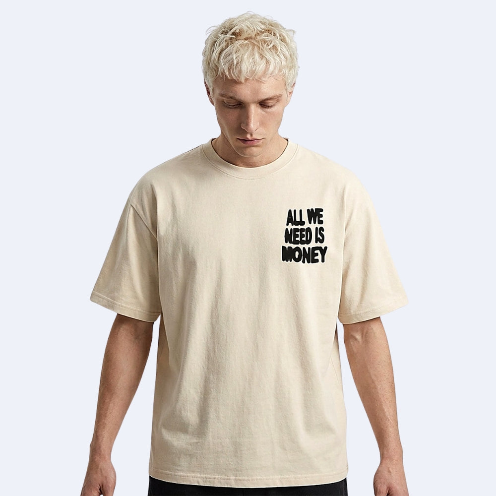 A man with short blonde hair wearing a beige crewneck t-shirt with the black text 'ALL WE NEED IS MONEY' printed on the left chest, set against a light gray background.