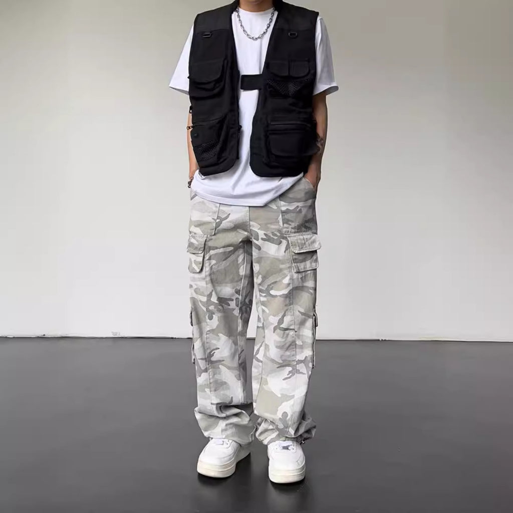 Person wearing a black vest, white t-shirt, camouflage pants, and white sneakers on a plain background