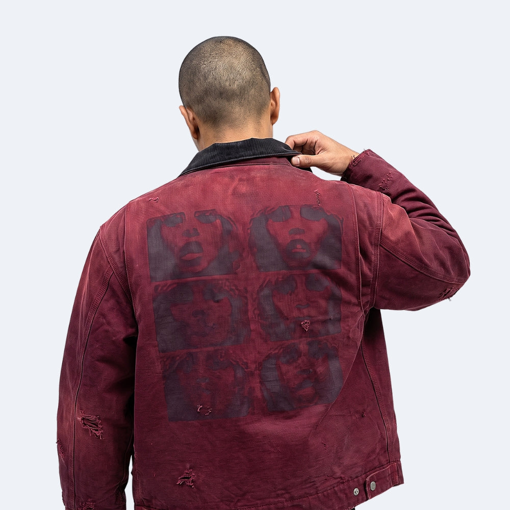 A man viewed from behind wearing a distressed maroon denim jacket with a large graphic print of four faces on the back, set against a plain light gray background.