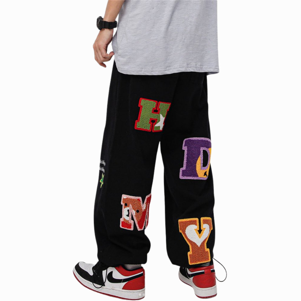 Black pants with colorful letter patches worn by a person on a white background