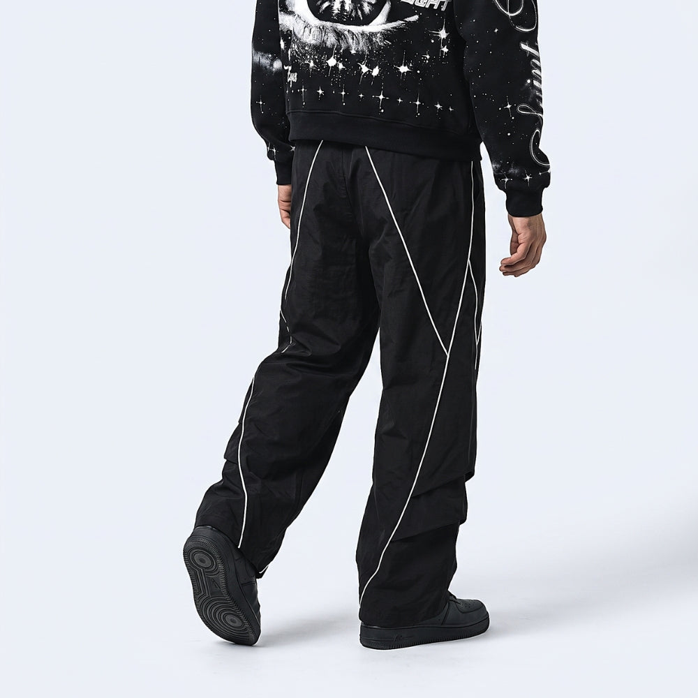 A model from the back wearing black wide-leg pants with white geometric line details, a black graphic sweatshirt, and black shoes, set against a light gray studio background.