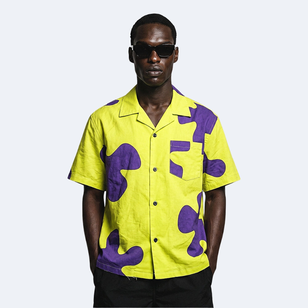 Yellow & Purple. A man wearing a bright yellow short-sleeve button-up shirt with bold purple abstract patches, styled with black sunglasses and black pants.