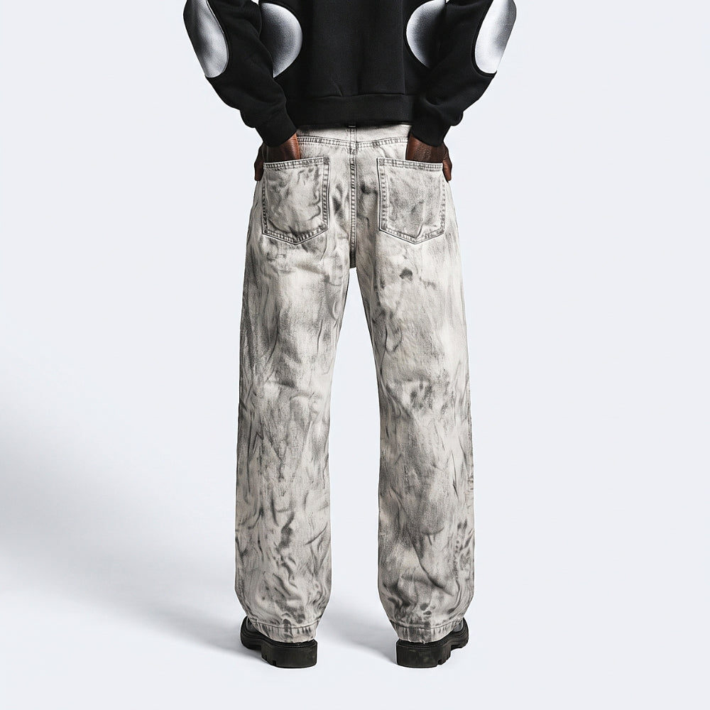 A person shown from the back wearing wide-leg jeans with a gray and white marble or tie-dye pattern, paired with a black hoodie featuring white circular patches on the sleeves and black shoes, against a light gray studio background.