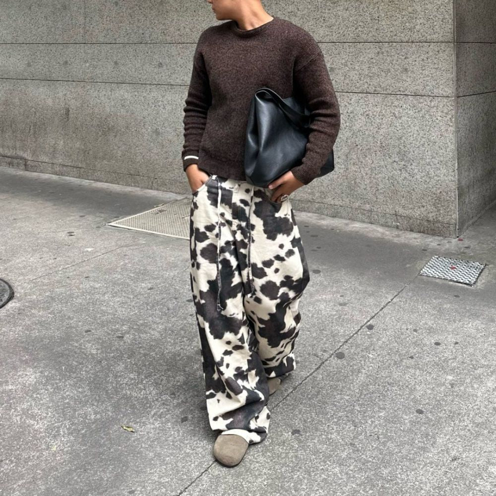 Person wearing a brown sweater and cow print pants standing on a sidewalk.