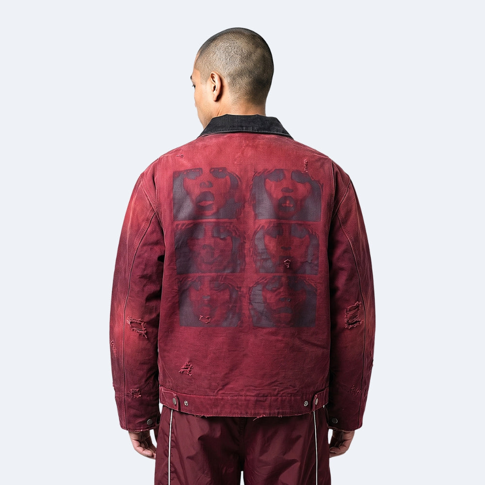 A man viewed from behind wearing a faded red distressed jacket with a large dark graphic print on the back, paired with matching dark red pants, against a light gray studio background.