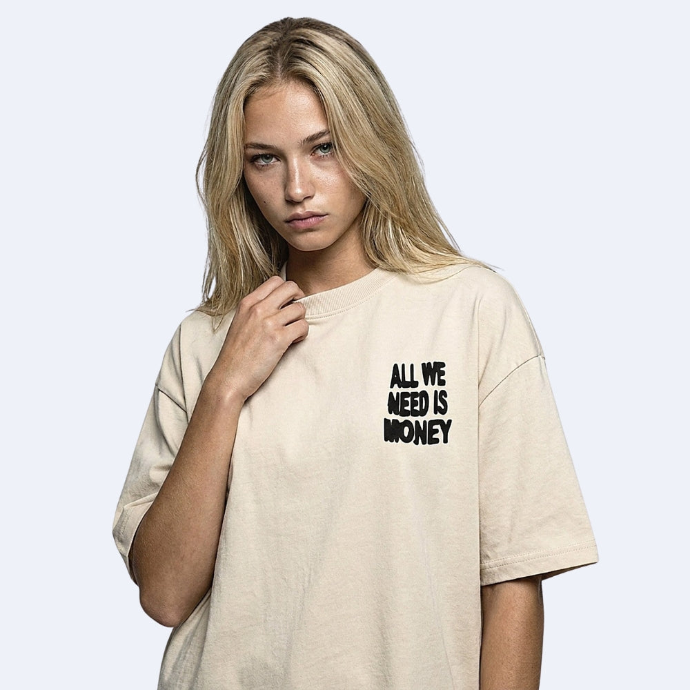 A blonde woman wearing an oversized beige t-shirt with the text 'ALL WE NEED IS MONEY' printed in black on the left chest, set against a light gray background.
