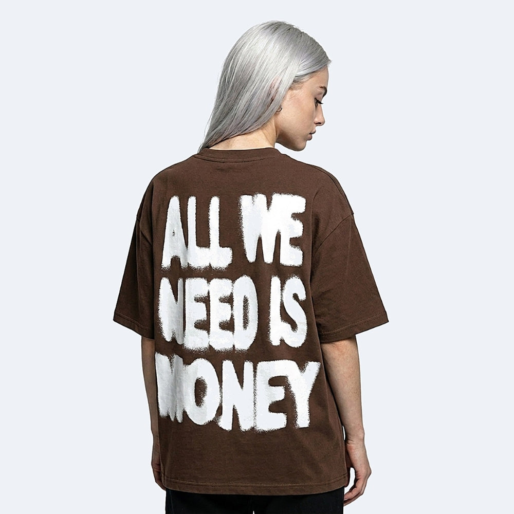 A person with silver hair shown from the back wearing an oversized brown T-shirt with large white distressed text reading 'ALL WE NEED IS MONEY' on a light gray studio background.