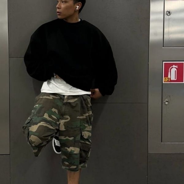 Person wearing a black sweater, white shirt, and camouflage pants leaning against a gray wall.