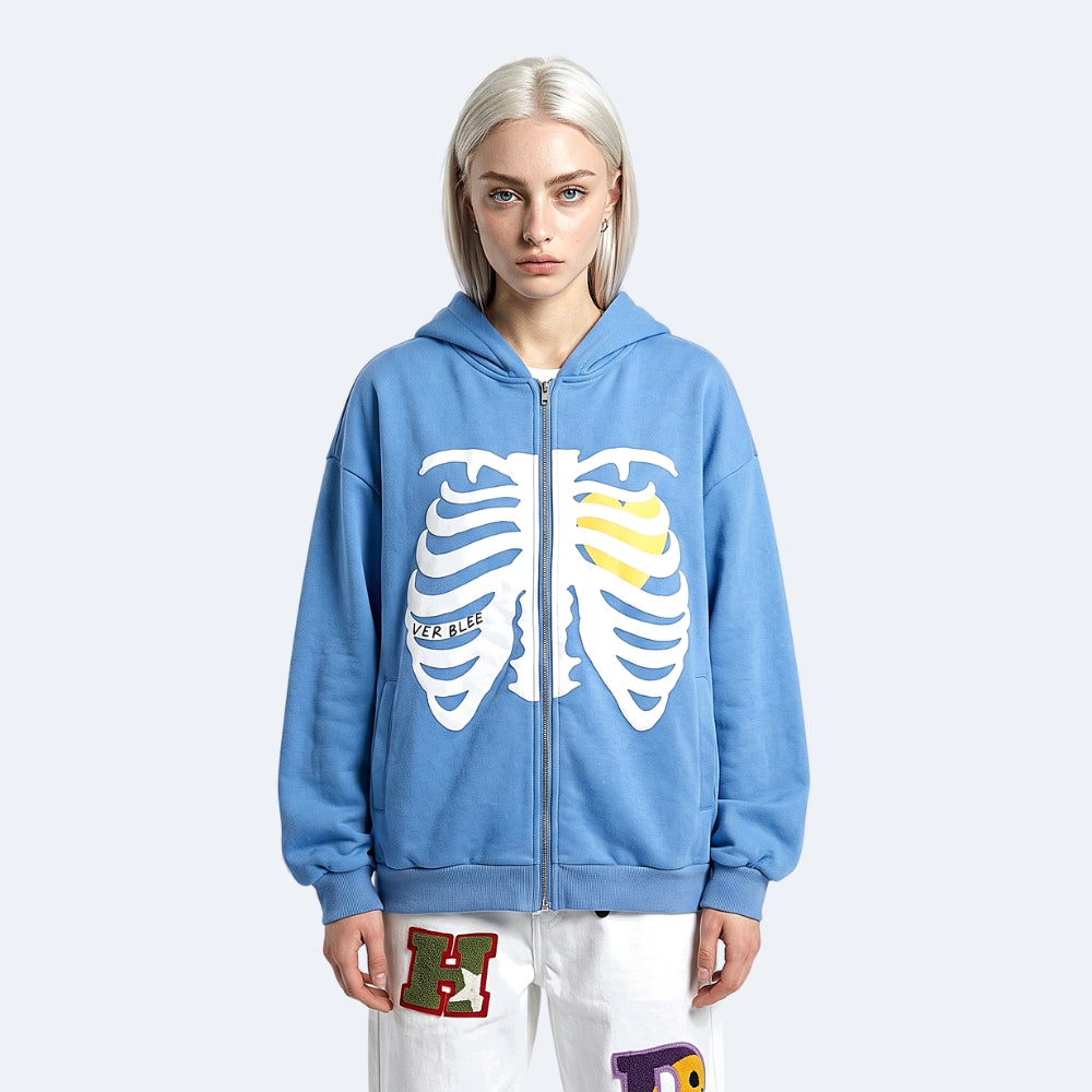 Blue. A model with platinum blonde hair wearing a light blue zip-up hoodie with a white rib cage graphic and a yellow accent, paired with white pants with colorful patches.