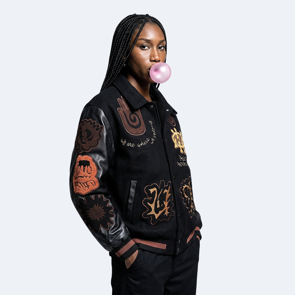 Black. A model wearing a black varsity-style jacket with leather sleeves and various colorful graphic patches.