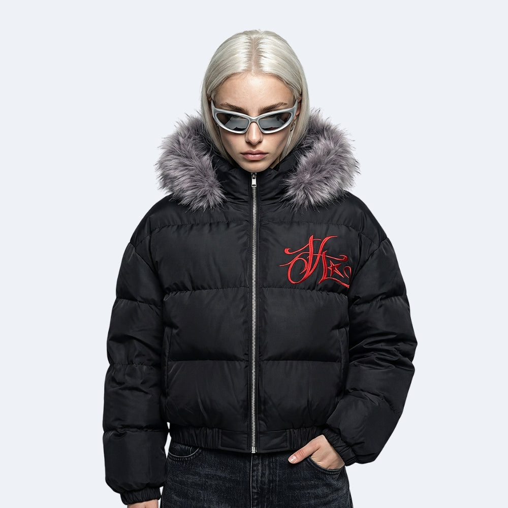 A model with platinum blonde hair and cat-eye sunglasses wearing a black puffer jacket with a gray faux-fur trimmed hood and a red embroidered logo on the chest, set against a light gray background.