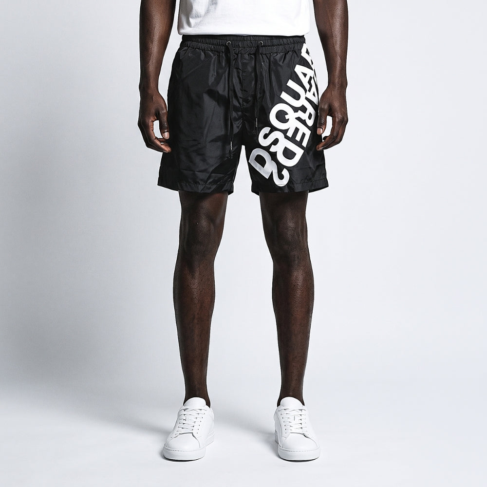 Black. A model wearing black athletic shorts with a bold white logo print down the side, paired with a white t-shirt and white sneakers.