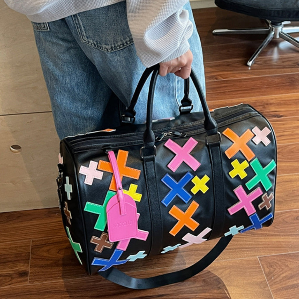 Black duffel bag with colorful geometric patterns held by a person on a wooden floor.