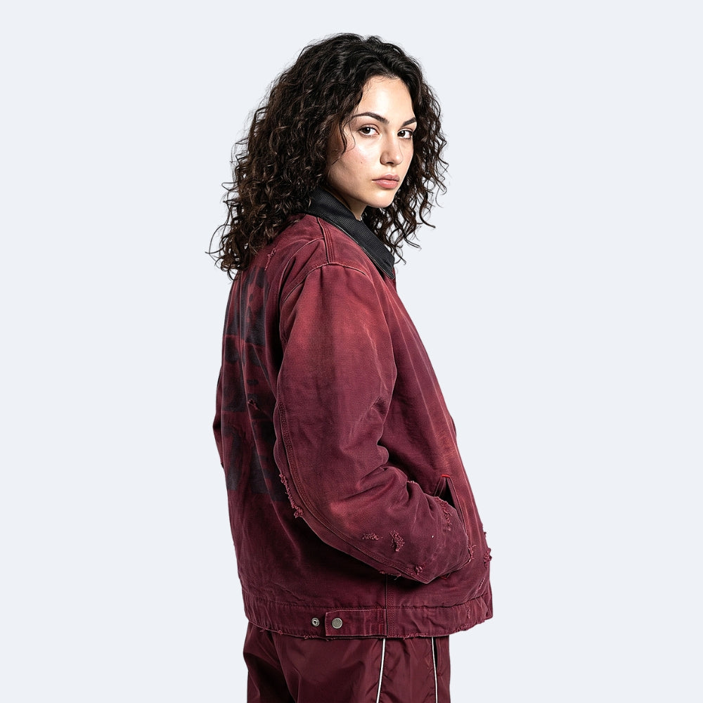 A model with curly dark hair is shown from the side/back wearing a distressed, faded burgundy utility jacket with a black collar and matching distressed pants against a light gray studio background.