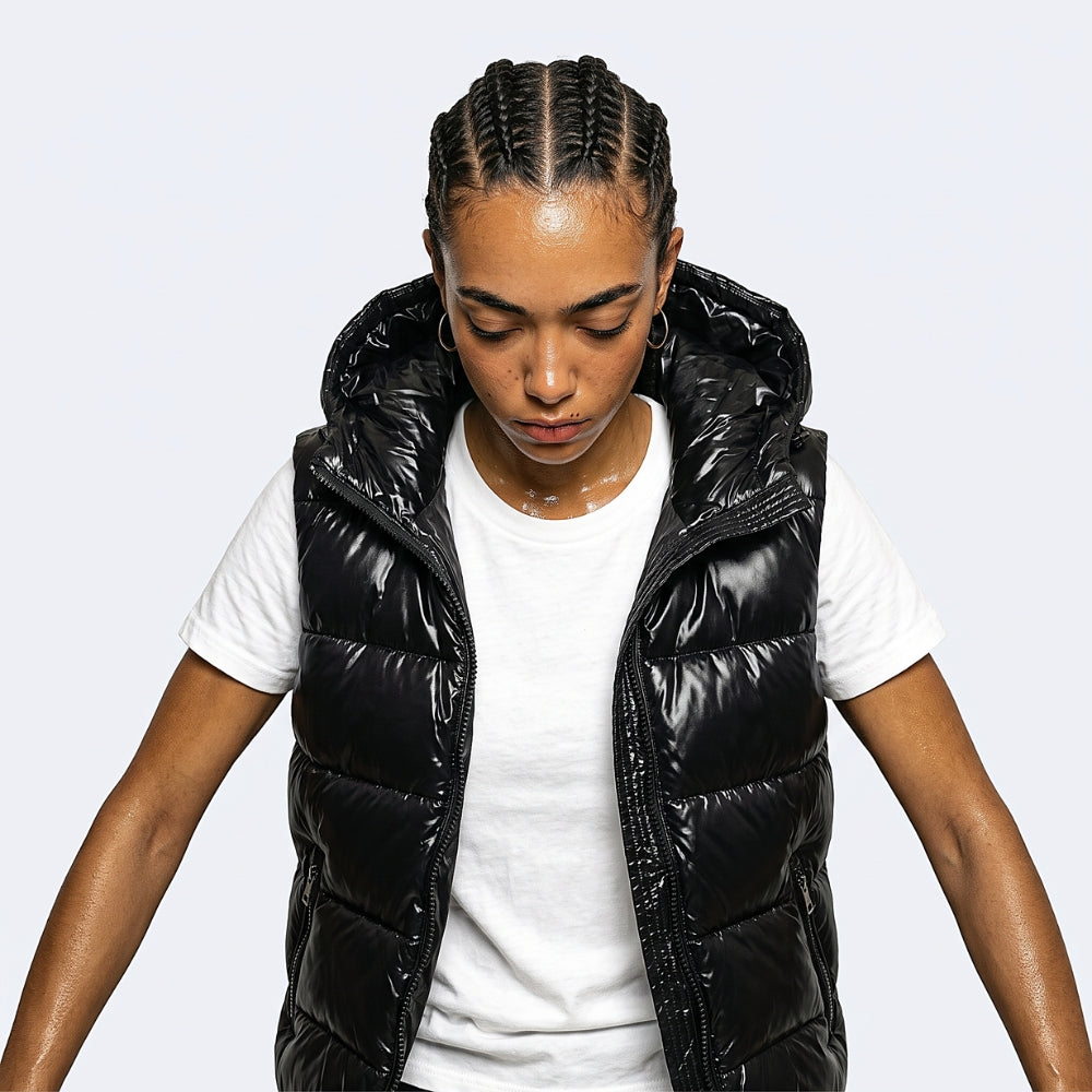 A woman wearing a shiny black puffer vest over a white t-shirt, looking down against a light gray background.