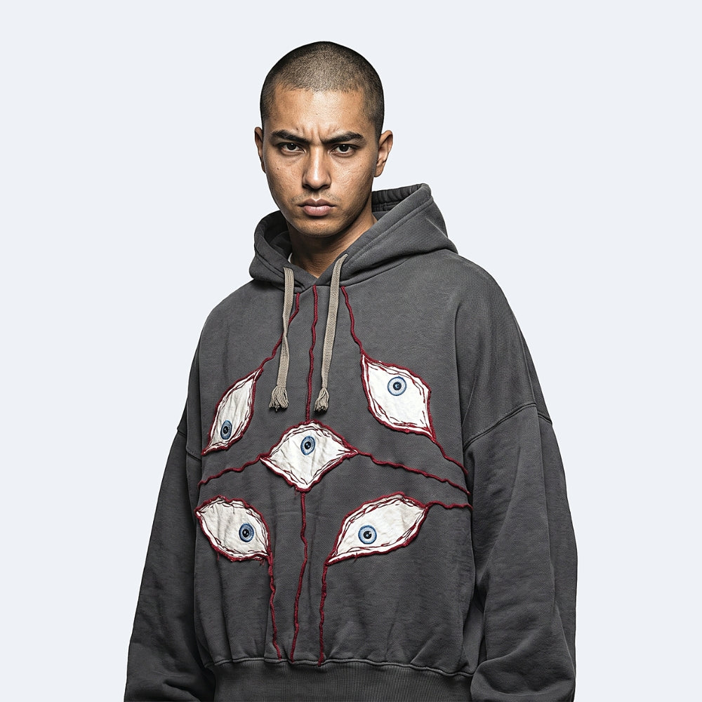 A man wearing a dark gray oversized hoodie with embroidered eyes on the front, posed against a light gray background.