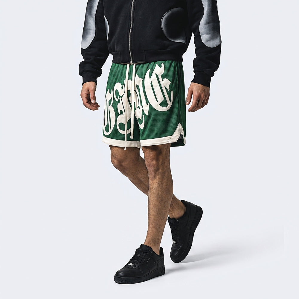 Green. A model wearing green athletic shorts with a large white gothic-style graphic print, paired with a black and gray zip-up hoodie and black sneakers.