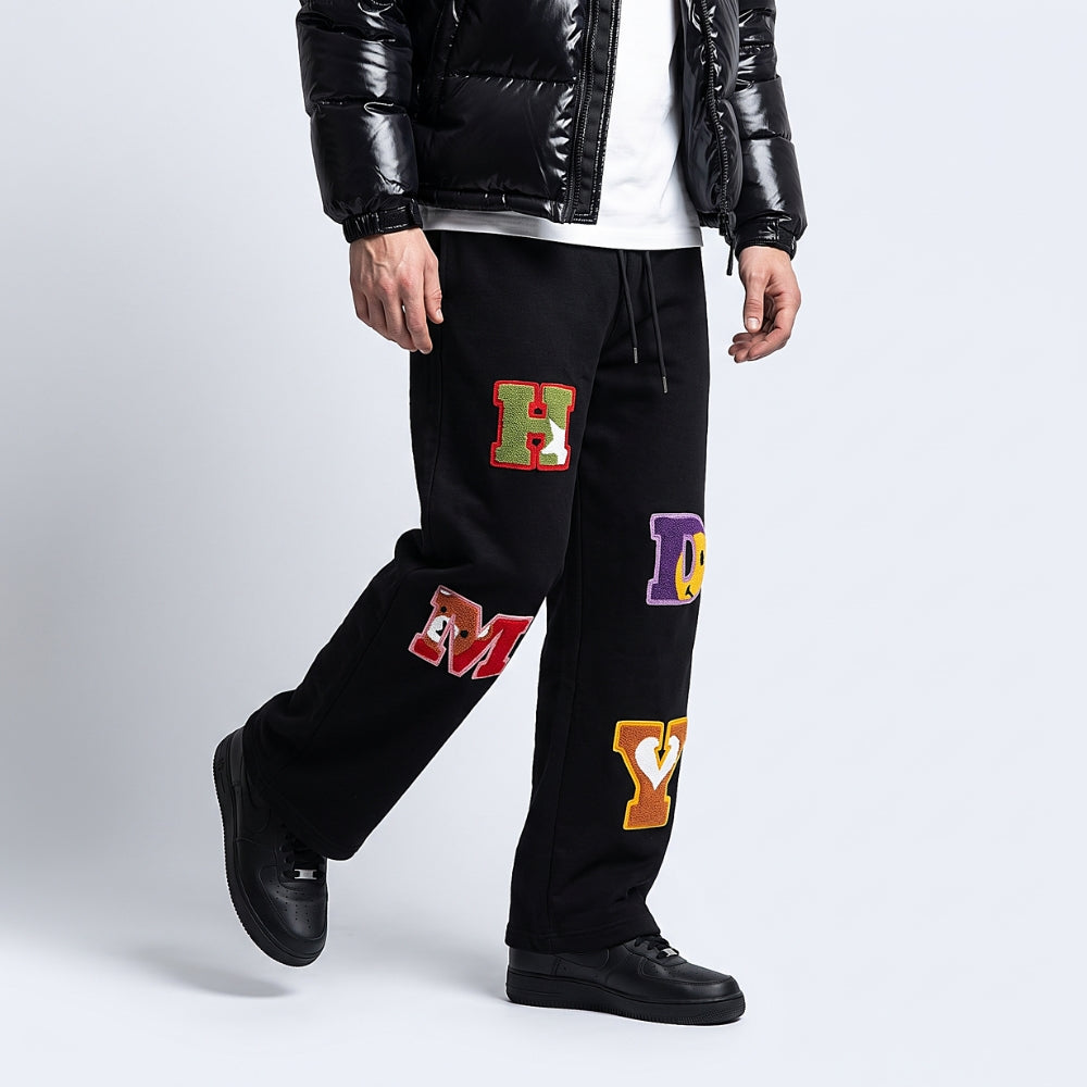Black. A person modeling black sweatpants with colorful varsity-style letter patches, paired with a black puffer jacket, white shirt, and black shoes.