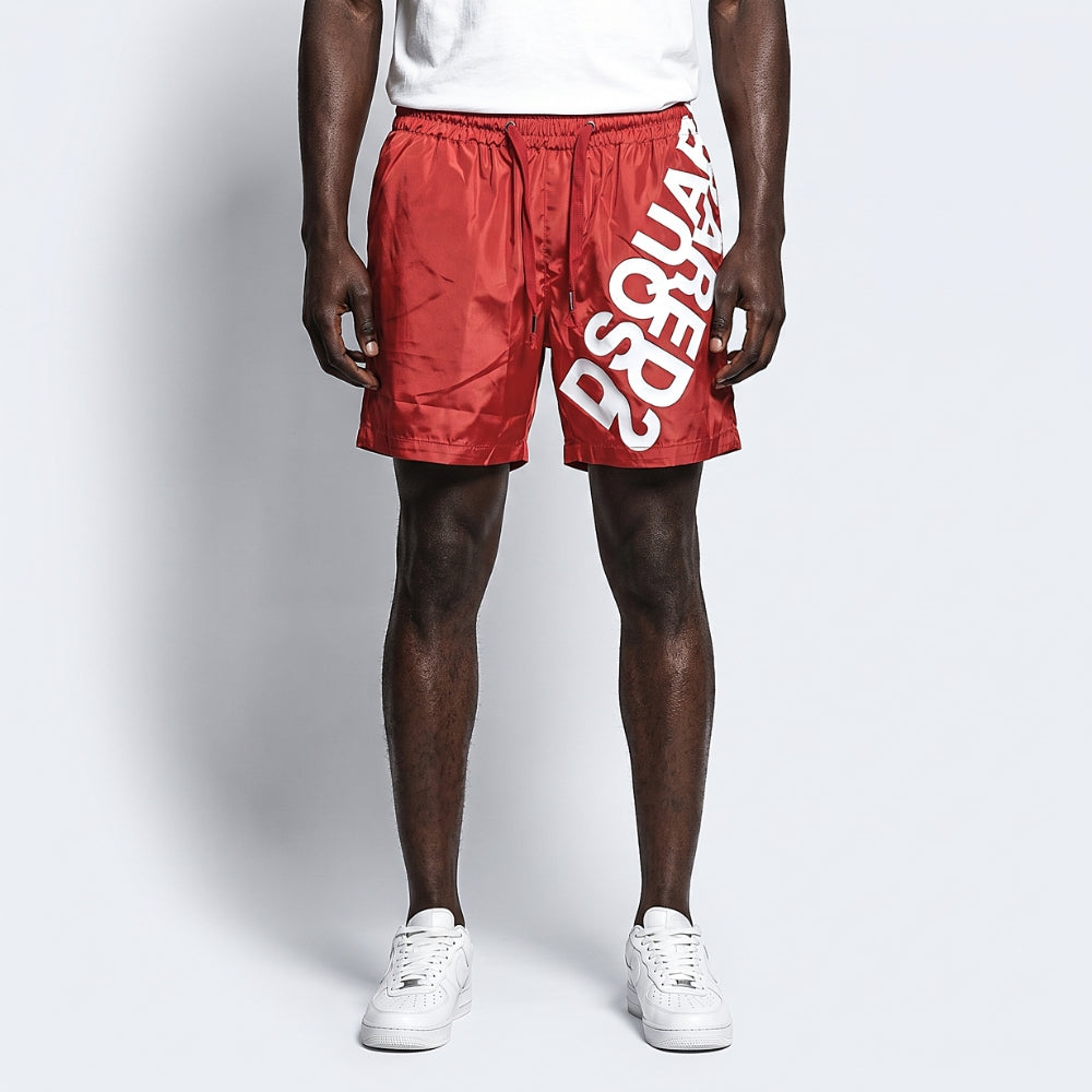Red. A model wearing red satin-like shorts with a large white diagonal logo print, paired with a white t-shirt and white sneakers.