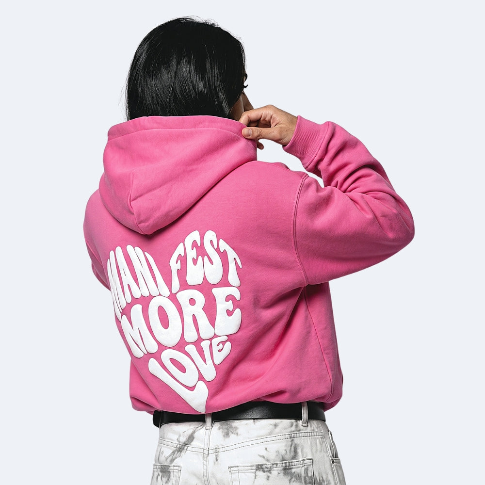 Pink. A person shown from the back wearing a bright pink cropped hoodie with large white text reading MANIFEST MORE LOVE, paired with light gray marbled jeans.