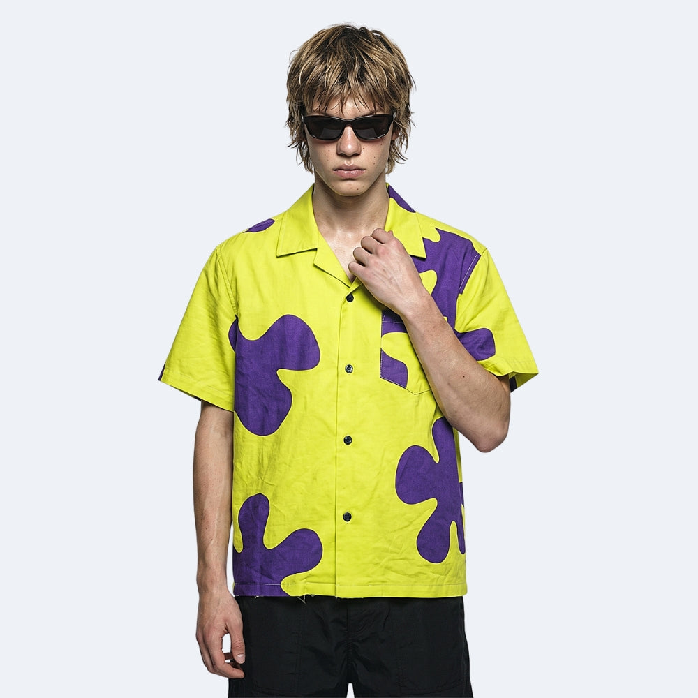 Yellow & Purple. A model wearing a bright yellow short-sleeve button-up shirt with a bold purple abstract pattern, styled with black sunglasses and black pants.
