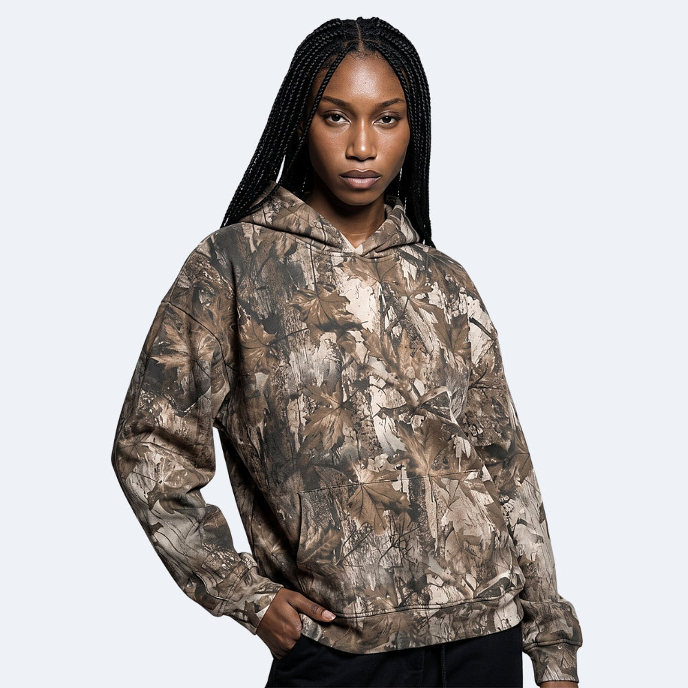 Oak. A woman modeling a camouflage hoodie with a realistic leaf and branch pattern in shades of brown, beige, and gray.