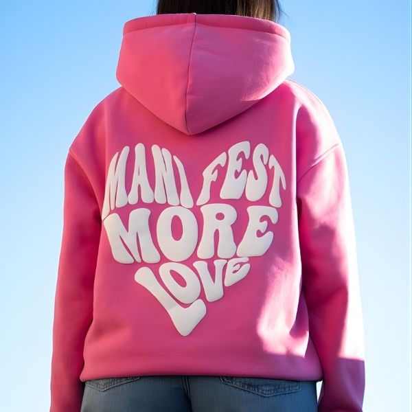 Pink hoodie with 'Manifest More Love' text on the back against a clear blue sky.