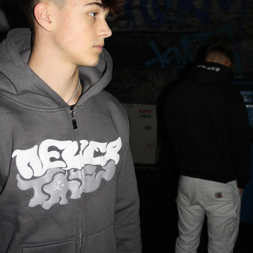 Person wearing a dark hoodie with a logo, standing indoors with another person in the background.
