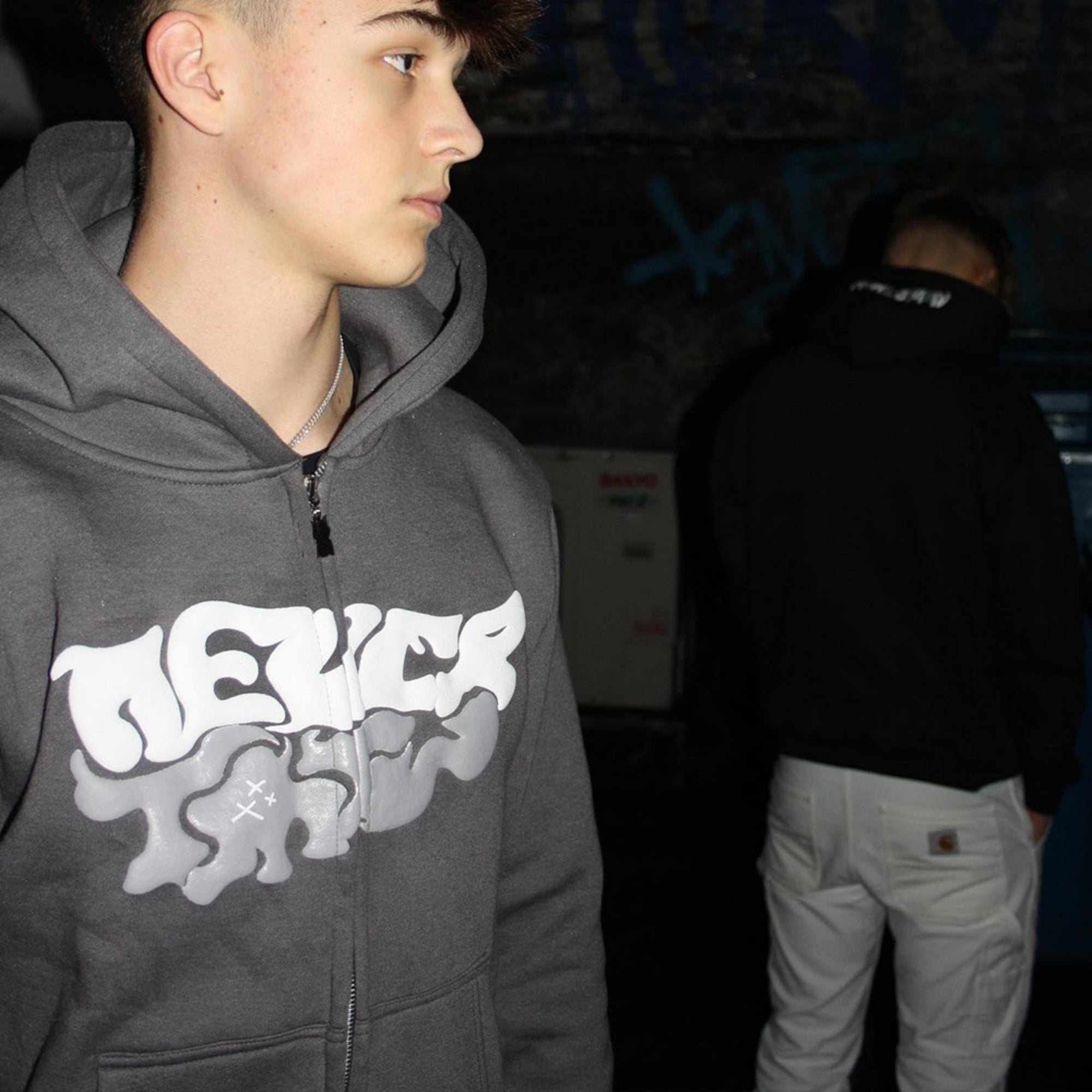 Person wearing a dark hoodie with a logo, standing indoors with another person in the background.