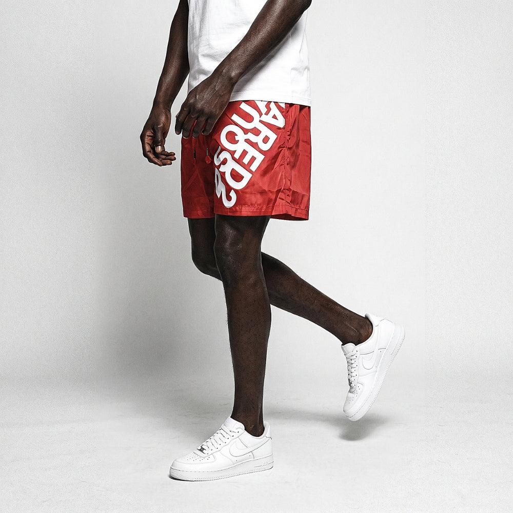 Red. A model wearing red shorts with a large white graphic on the side, paired with a white t-shirt and white sneakers.