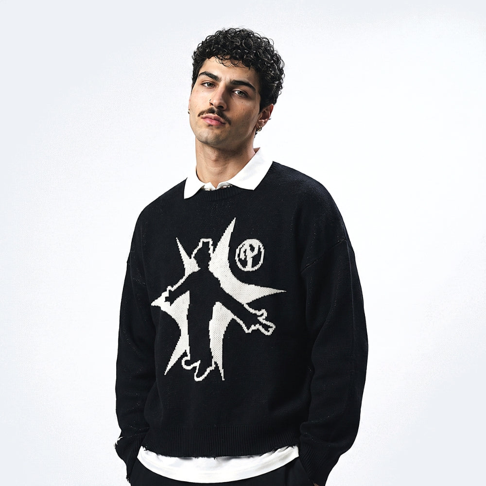 A man wearing a black knit sweater with a white graphic design on the front, layered over a white collared shirt, against a plain white studio background.