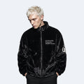A man with platinum blonde hair wearing a black plush fleece jacket with white text on the chest and a white circular graphic on the sleeve, posed against a light gray background.