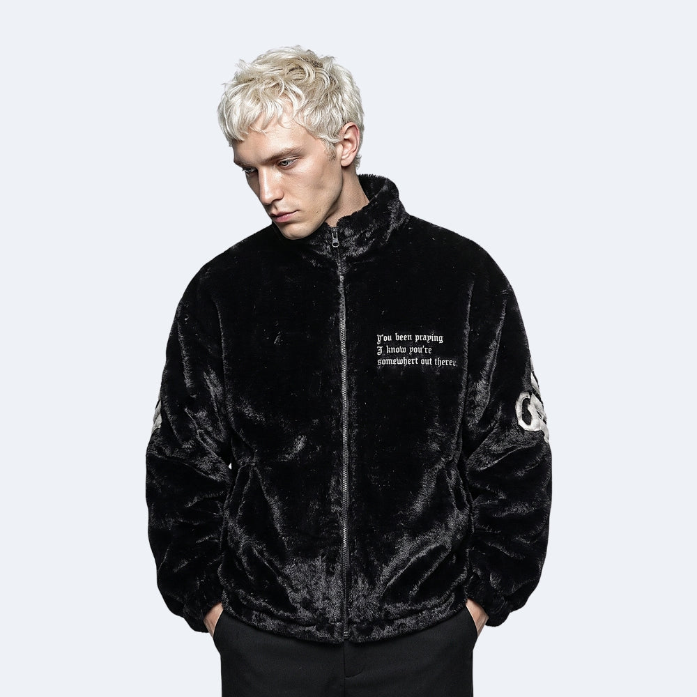 A man with platinum blonde hair wearing a black plush fleece jacket with white text on the chest and a white circular graphic on the sleeve, posed against a light gray background.